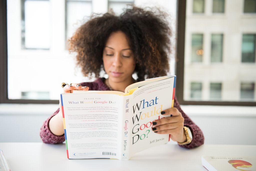 A woman reading 'What Would Google Do?' at a desk by a window in a modern office.